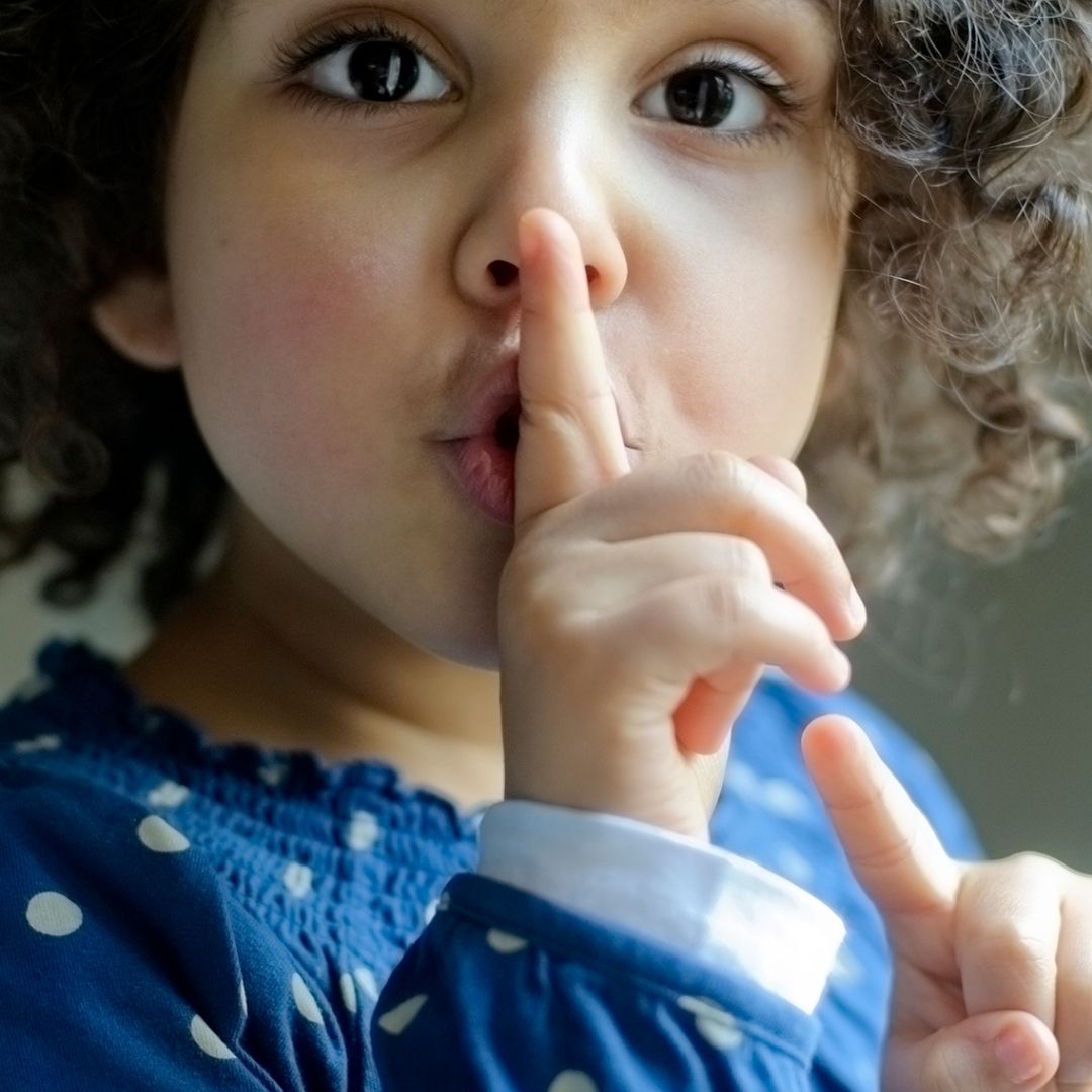 A little child holding one finger up to their lips as if telling someone to be quiet.