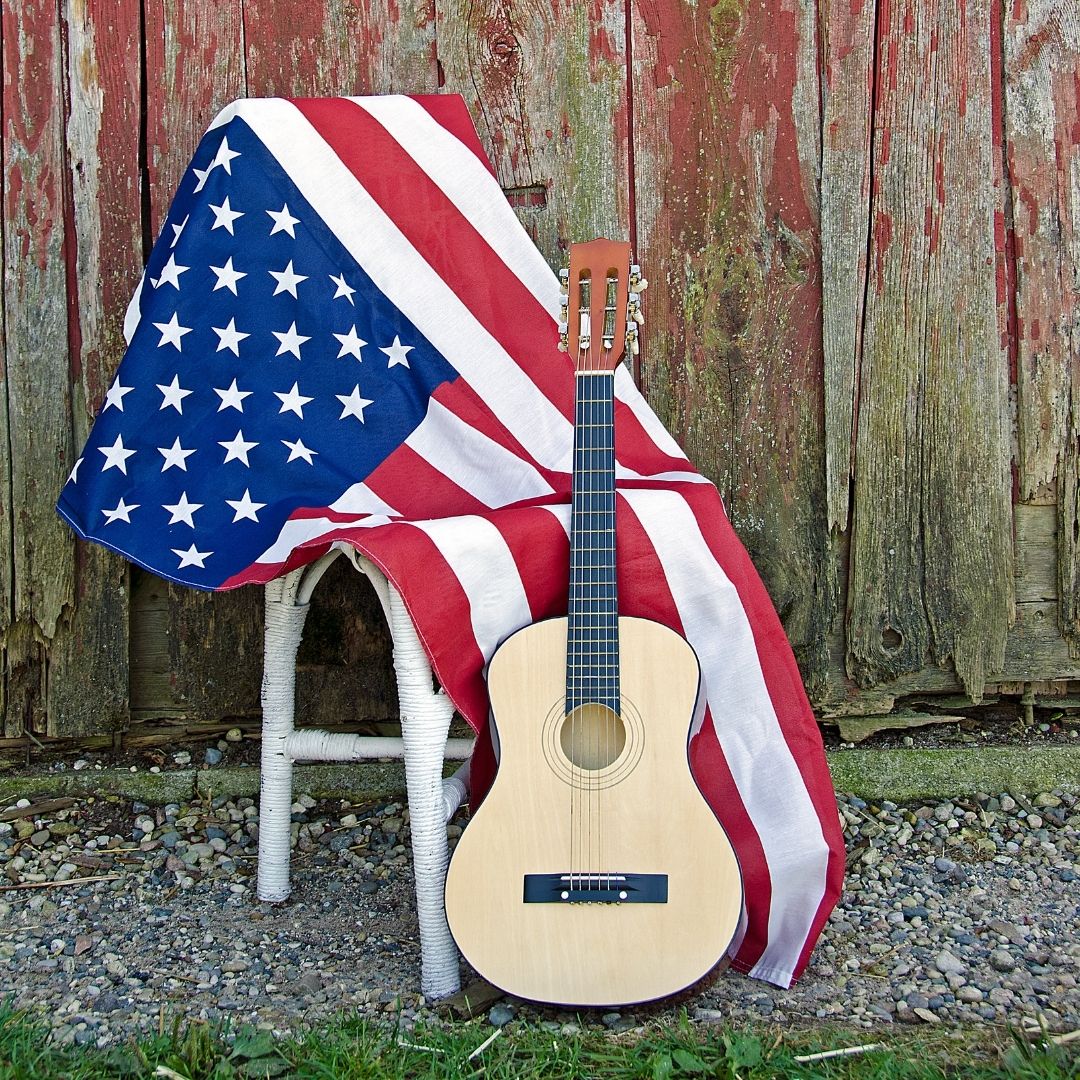 An acoustic guitar leaning against a white chair that has an American red, white, and blue flag draped over it.