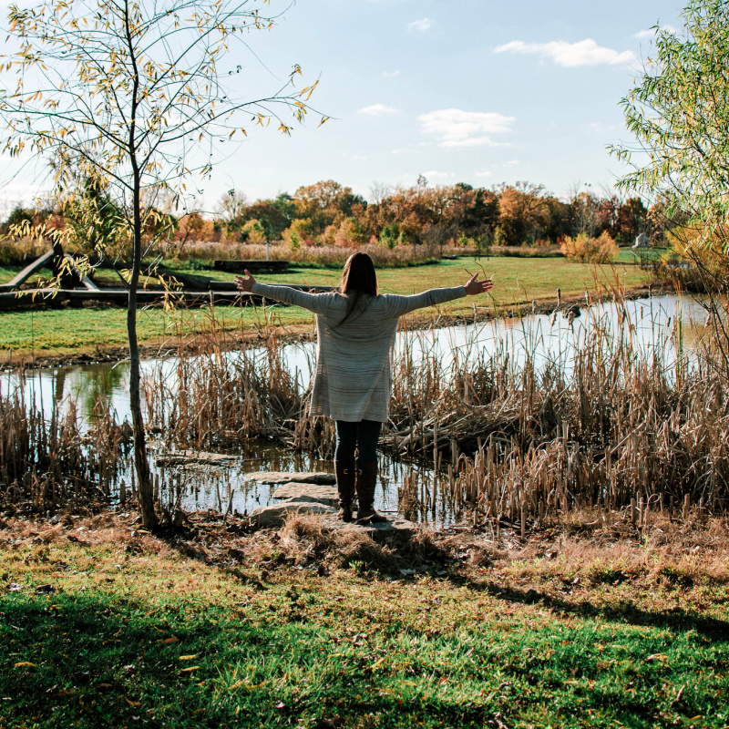 Brittany standing on the edge of a pond with her arms out stretched, facing the sun.