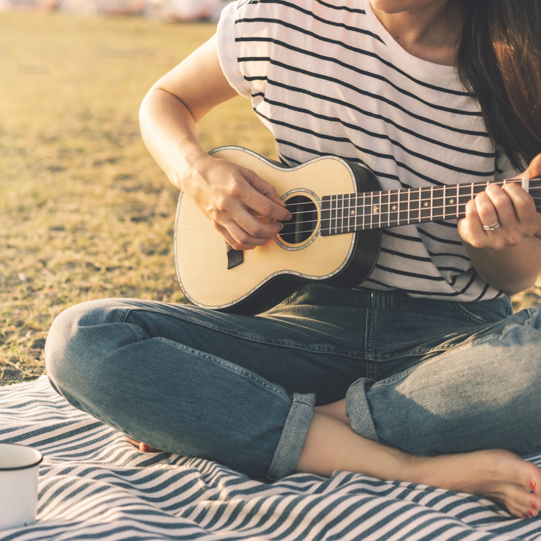A person in a striped shirt and jeans sits on a striped blanket, strumming a ukulele outdoors.