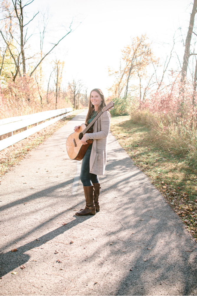 Living Music LLC owner and music therapist Brittany Scheer holds a guitar while walking along a pathway.